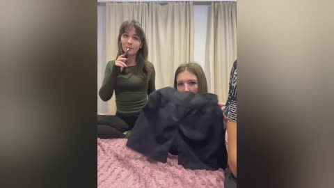 Video of a young woman with long brown hair in a green top, holding a phone while another woman with long blonde hair, wearing a black top, lies on a pink bed, covered by a black blanket.