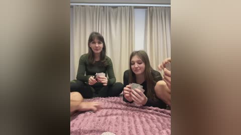 A video captures two women in a cozy room with beige curtains. One woman, with long brown hair, wears a green top; the other, with straight brown hair, dons a dark sweater. They sit on a pink blanket, holding small objects, creating a warm, intimate scene.