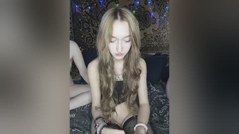 Video of a slender, fair-skinned, long-haired young woman with light makeup, wearing a black crop top, sitting on a bed with dark bedding, surrounded by colorful string lights and patterned tapestry in the background.
