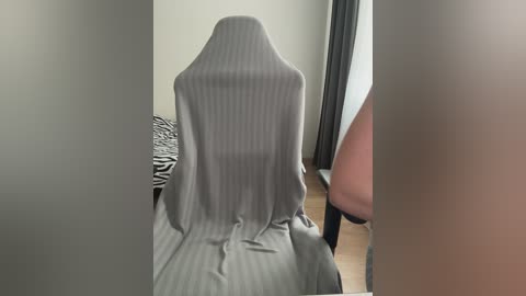 Video of a grey-striped cloth draped over a wooden chair, partially obscuring a person's lower body in a room with light-colored walls and zebra-patterned bedding.