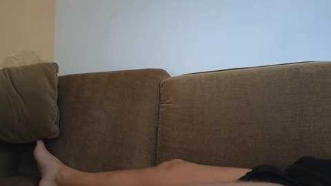 Video of a beige sectional sofa against a plain, light-colored wall. A person's bare, tan legs are visible in the foreground, suggesting a casual, relaxed setting.