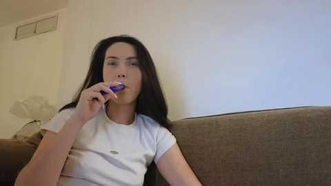 Video of a young woman with long black hair, fair skin, and small breasts, wearing a white t-shirt, brushing her teeth with a purple toothbrush, sitting on a brown couch in a minimalist living room.