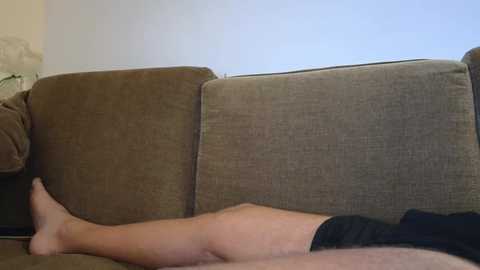 A video showing a person lying on a beige and brown fabric couch with a beige wall in the background. The person is barefoot, wearing black shorts, and their legs are crossed.