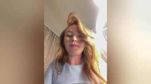 A video of a fair-skinned woman with long, wavy, red hair, wearing a light blue shirt, smiling with closed eyes, standing in a softly lit room with beige walls and a window.