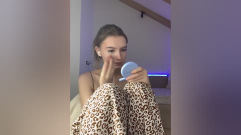 Video of a young woman with light skin, brown hair, and minimal makeup, wearing leopard-print pajamas, sitting on a bed, applying makeup with a light blue compact mirror.