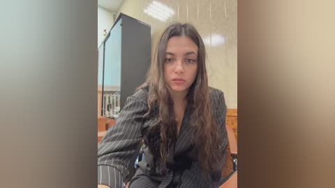 Video of a young woman with long brown hair, wearing a dark pinstriped suit, sitting at a wooden desk in a classroom with beige walls and fluorescent lighting.