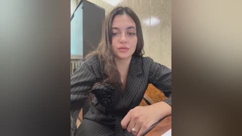 Video of a young woman with long brown hair, wearing a black pinstriped suit, seated on a wooden chair against a beige wall, in a dimly lit room.