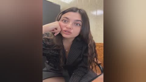 Video of a young woman with long, dark hair, wearing a black pinstripe blazer and a fur-trimmed jacket, leaning against a wall in a dimly lit room.