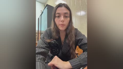 Video of a young woman with long brown hair, wearing a dark pinstriped dress, seated in a dimly lit room with a beige wall and wooden floor.