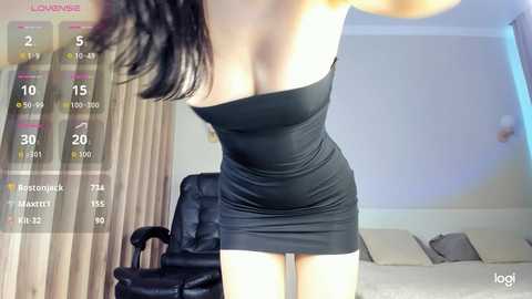 Video of a woman in a tight black dress standing in a modern, well-lit living room with beige curtains and a black leather armchair.