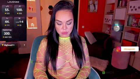 A video of a curvaceous Latina woman with long black hair, wearing a neon green fishnet bodysuit, sitting in a dimly-lit room, with a live cam overlay on the left.