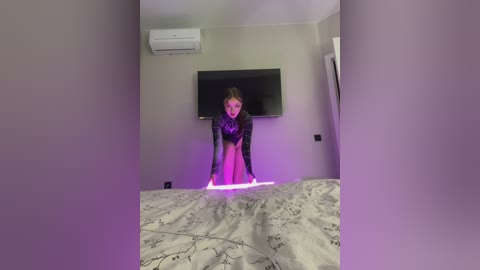 A video of a woman in a black, sequined leotard standing on a bed with a gray floral sheet, illuminated by purple light. She has fair skin, blonde hair, and stands in a modern, beige-walled bedroom with a mounted flat-screen TV and air conditioner.