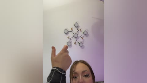 Video of a young woman with light skin, brown hair, wearing a denim jacket, pointing at a white wall with a decorative, circular metal art piece in the center.