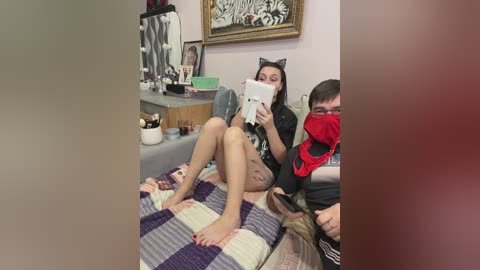 A video shows a woman with cat ears reading a magazine, a man in a mask, and a room with framed art, cluttered shelves, and a checkered blanket.