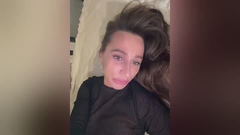 A video of a light-skinned woman with long, voluminous brown hair, wearing a black mesh top, lying on a white bed with a beige wall behind. She has a sultry expression.