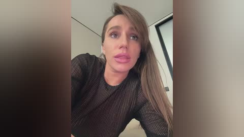 A video of a Caucasian woman with long, straight brown hair, wearing a sheer black top, with a surprised expression, indoors with a white wall and window in the background.