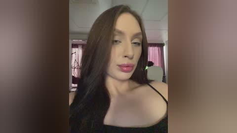 Video of a young Caucasian woman with long, straight dark hair and light skin, wearing a black off-shoulder top, standing indoors with pink curtains and a bed visible in the background.