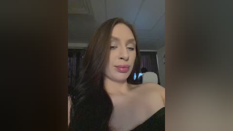 Video of a young woman with fair skin, long brown hair, and a black off-shoulder dress. She has a neutral expression, looking slightly to the left. Background includes a dimly lit room with a white ceiling and dark curtains.