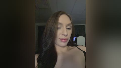 A video of a young woman with long, dark hair, fair skin, and wearing a black strapless top, taken indoors with dim lighting. She has a soft, serene expression.