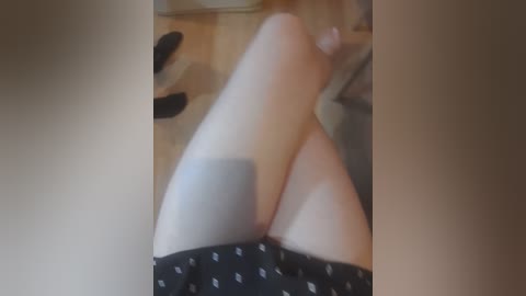 Video of a light-skinned woman's legs, covered in black shorts, lying on a wooden floor. Shoes and a white object are visible in the background.