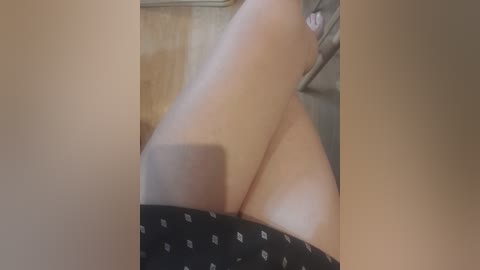 Video of a close-up of a woman's thighs and buttocks, wearing black shorts with white diamond patterns. Her skin is fair, and the background shows a wooden wall and a blurry part of a person's torso.
