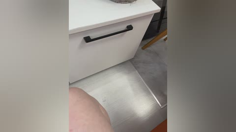 Video of a person's hand holding a black, rectangular object, standing in a room with a white cabinet and grey floor, likely a kitchen or bathroom.
