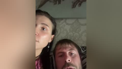 Video of two people, a woman with long dark hair and a man with a beard, sharing a close, intimate moment. The woman's face is partially hidden, while the man's expression is intense. Background shows a floral patterned wallpaper.