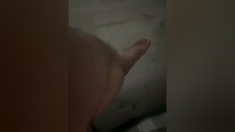 A dimly-lit, close-up video of a barefoot man's foot and lower leg, with his toenails visible, standing on a concrete floor. The image is grainy and slightly blurred.