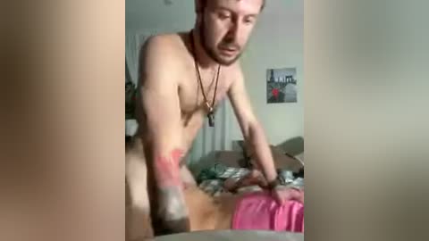 Video of a muscular, light-skinned, bearded man with short hair, wearing a necklace, standing naked in a bedroom with messy bedding, walls, and a small picture on the wall.