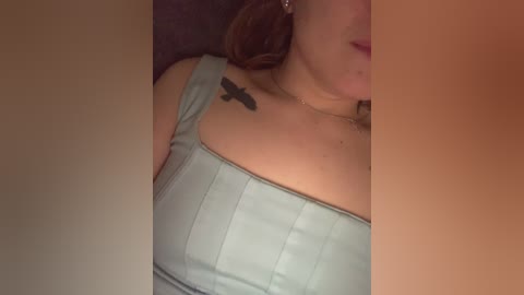 Video of a woman with fair skin, wearing a light blue, ribbed tank top, showcasing a small black bird tattoo on her collarbone. Her lips are painted a soft pink.