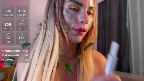 Video of a blonde woman with green makeup on her face, holding a white lipstick, in a dimly lit room.