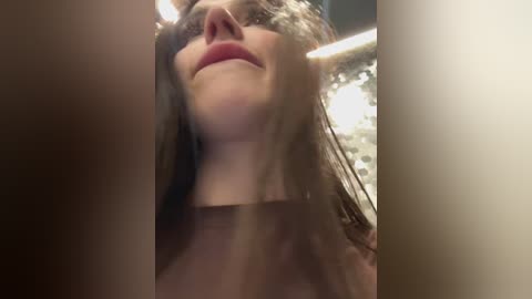 A close-up video of a smiling woman with long brown hair and fair skin, wearing a brown top. Background is blurred, featuring warm lighting and green foliage.