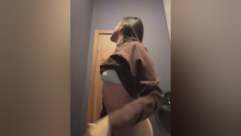 A video of a pregnant woman in a dimly lit hallway, wearing a brown jacket over a white bra, her belly prominent, with a wooden door visible behind her.