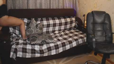 A video shows a woman arranging a plaid-patterned bedspread on a dark wooden couch with decorative pillows. The background features a beige wall with floral wallpaper and a black leather office chair.