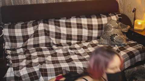 Video of a cozy, dimly lit bedroom with a plaid-covered bed, a candle-lit nightstand, and a person with purple hair, blurred in the foreground.