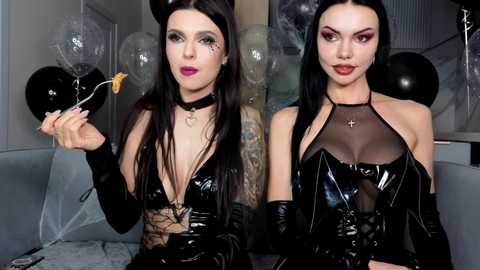 Video of two women in black latex dresses and gloves, one holding a small doll, both with long dark hair, tattoos, and dark makeup. Background includes black balloons and a gray couch.