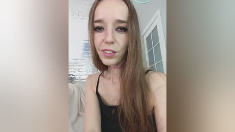 A video of a young woman with long brown hair, pale skin, and a thin build, wearing a black tank top and a pink choker. Background features a white door and a white wall with abstract art.