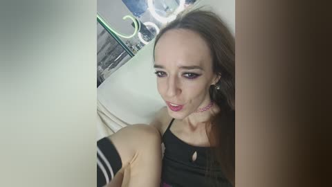 Video of a slender, pale-skinned woman with long brown hair, wearing black makeup, a black top, and pink choker, sitting on a white couch, surrounded by mirrored walls and neon lights.