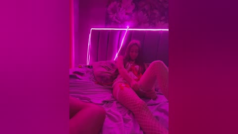 Video of a woman in fishnet stockings and a lacy bra, lying on a bed with a floral-patterned duvet. Neon pink lights illuminate the room, creating a vibrant, sensual atmosphere.