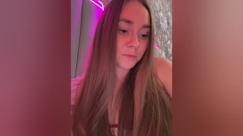 Video of a young Caucasian woman with long, straight brown hair, wearing a white top, standing in a dimly lit room with pink and purple lighting.