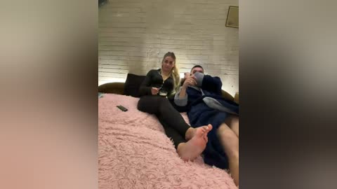 Video of a man and woman on a pink shag carpeted bed in a dimly lit room with white brick walls. The man is holding a phone, the woman is looking at him.