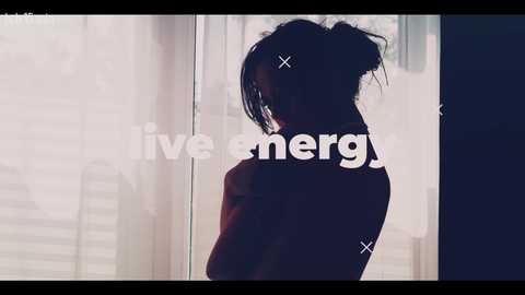 Video of a young woman with dark hair tied in a messy bun, silhouetted against a window with sheer curtains, revealing an urban skyline. She wears a red sweater, looking introspective.