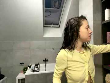 Video of a young woman with wet, dark hair, wearing a yellow cardigan, standing in a modern, white-tiled bathroom with a skylight, black shelves, and a white bathtub.