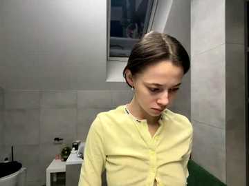 Video of a young woman with light skin and straight, shoulder-length brown hair, wearing a light yellow blouse, in a modern, white-tiled bathroom with a small window and toiletries on the sink.