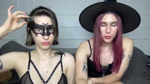 Video of two women with pale skin, one with short brown hair and a black lace mask, the other with long red hair and a black witch hat, both wearing black lace lingerie.