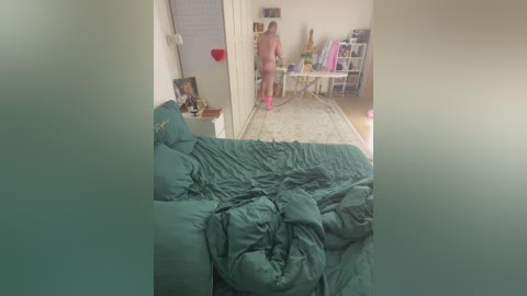 Video of a messy, teal-tinted bedroom with a woman in pink underwear standing at the doorway. Unmade bed with rumpled sheets and scattered clothes. Background shows a cluttered room with a white desk, shelves, and a white door.
