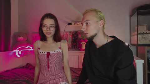 Video of a young woman with glasses and a red-and-white checkered dress, seated next to a man with short blonde hair and a black shirt, in a dimly lit, pink-lit room with a fish tank and a neon sign.