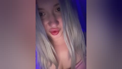 Video of a young woman with fair skin, long platinum blonde hair, and full lips. She's wearing a pink top, posing seductively with her head slightly tilted, against a purple-lit background.