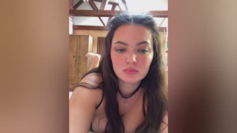 A video of a young woman with long dark hair, fair skin, and full lips, wearing a black choker necklace, lying on her stomach in a rustic room with wooden walls and beams.