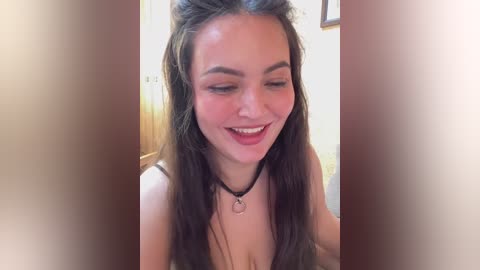 Video of a smiling young woman with long brown hair, wearing a black choker necklace, in a bright room with wooden furniture and beige walls.